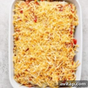 Creamy Cottage Cheese Taco Lasagna 12 A white baking dish filled with an unbaked casserole topped with shredded cheese on a light-colored surface.