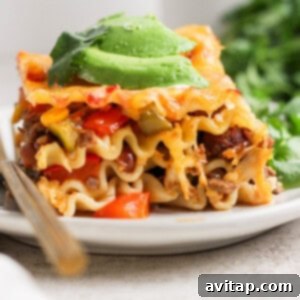 A close-up of a perfectly baked and portioned slice of Whipped Cottage Cheese Taco Lasagna, topped with creamy avocado slices, on a white plate with a fork.