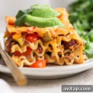 Creamy Cottage Cheese Taco Lasagna 14 A close-up of a slice of lasagna topped with avocado slices on a white plate, with a fork beside it and green herbs in the background.