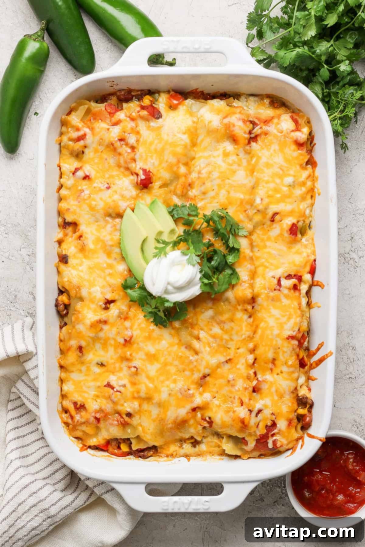 A baked casserole topped with melted cheese, avocado slices, sour cream, and cilantro, presented in a white baking dish. Fresh jalapeños, cilantro, and a dish of red sauce are beside it, ready to be served alongside our Whipped Cottage Cheese Taco Lasagna.