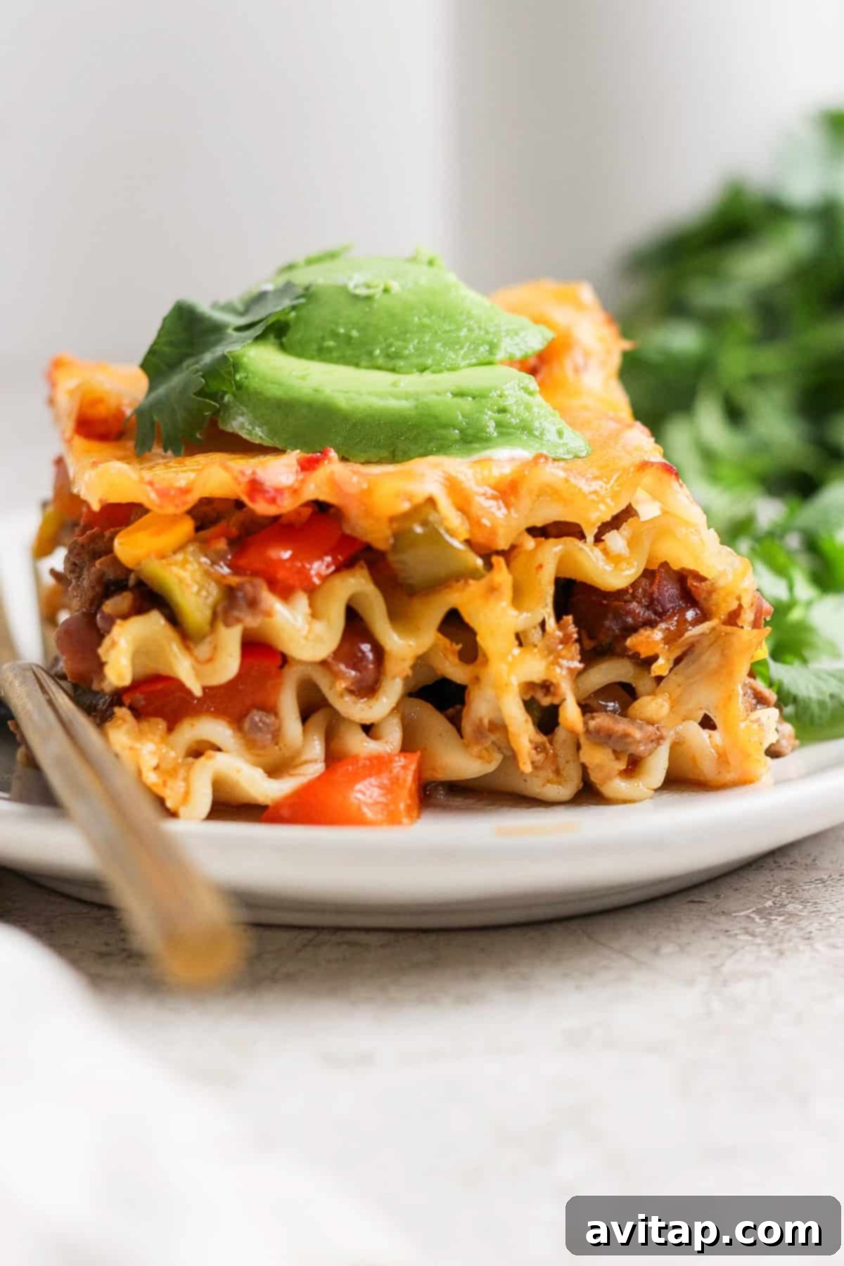 A close-up of a perfectly portioned slice of Whipped Cottage Cheese Taco Lasagna topped with fresh avocado slices, served on a white plate with a fork beside it, and green herbs in the background.