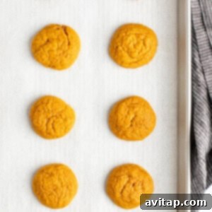 Eight freshly baked pumpkin cookies cooling on a parchment-lined baking sheet, with a gray cloth visible on the right side, signifying the completion of the baking step.