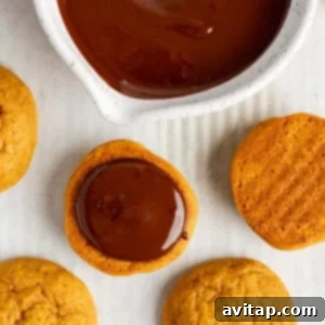 Chocolate Kissed Pumpkin Cookies 12 A pumpkin cookie being carefully dipped into a bowl of smooth, melted chocolate. Additional cookies are waiting nearby on a white surface.