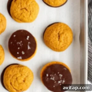 Chocolate Kissed Pumpkin Cookies 13 A baking tray with parchment paper holds nine round orange cookies. Some are plain, others are half-dipped in chocolate and sprinkled with sea salt.