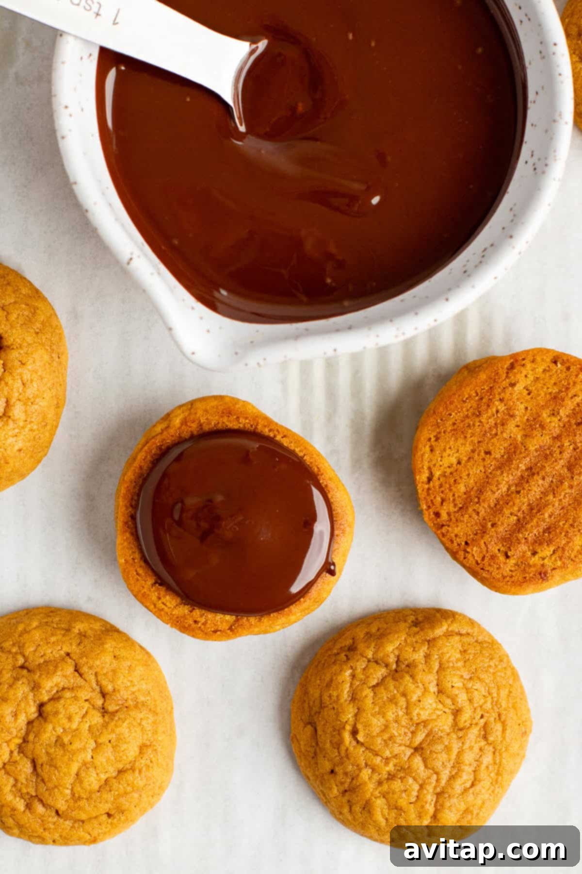 Freshly baked pumpkin cookies being carefully dipped into a bowl of smooth, melted chocolate. Additional plain cookies are neatly arranged on a white surface nearby, awaiting their delicious coating, creating an inviting visual of the baking process.