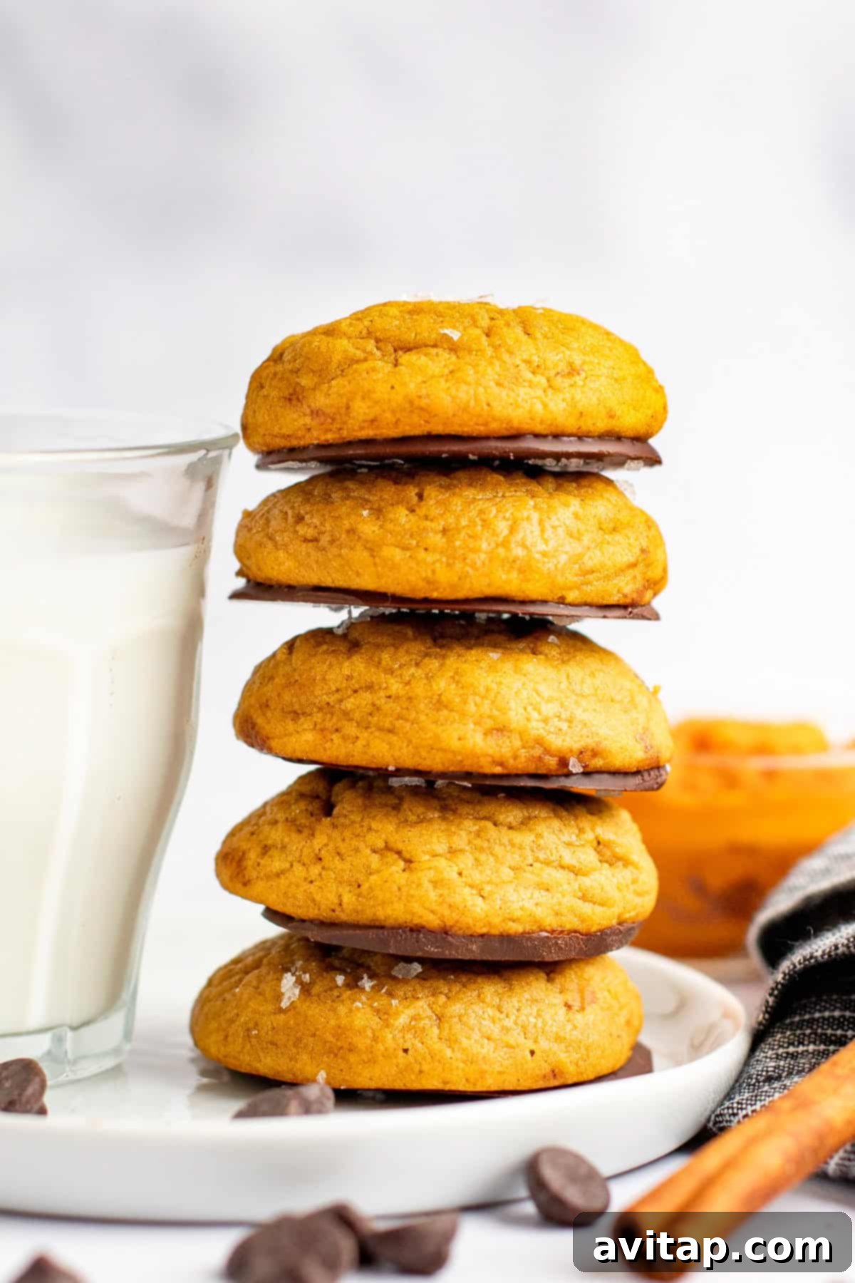 Chocolate Kissed Pumpkin Cookies 6 A stack of five golden-orange pumpkin cookies on a white plate, with a glass of milk and a folded cloth napkin nearby. Chocolate chips are scattered around, emphasizing the chocolate component of the dessert.