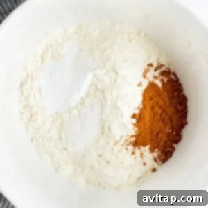 Chocolate Kissed Pumpkin Cookies 8 A white bowl containing flour, cinnamon, and baking powder, with a striped cloth partially visible in the background.