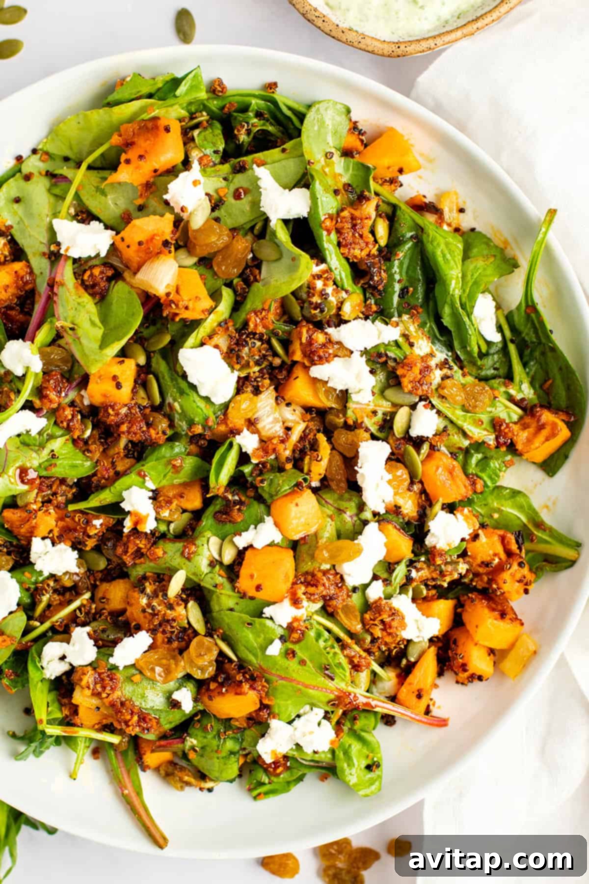 A plate of salad with mixed greens, roasted butternut squash, crumbled feta cheese, quinoa, pumpkin seeds, and golden raisins.