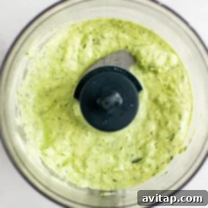 A bowl of green and white sauce sits inside a food processor with a blade in the center.