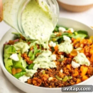 A hand pouring green herb dressing from a jar onto a bowl containing quinoa, roasted vegetables, greens, seeds, and crumbled cheese with another bowl of roasted vegetables in the background.