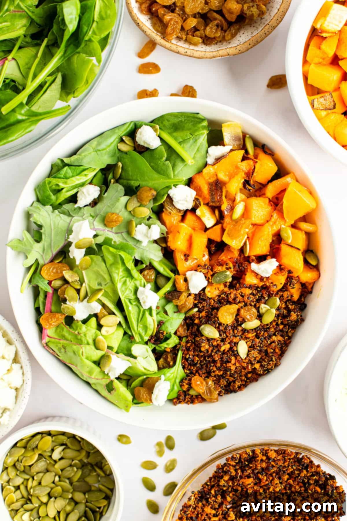 A bowl of salad with spinach, roasted butternut squash, quinoa, pumpkin seeds, and goat cheese crumbles, surrounded by small bowls of salad ingredients, including raisins and seeds.