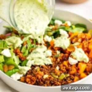 A close-up of a salad in a white bowl with quinoa, mixed greens, pumpkin seeds, raisins, and crumbled cheese, being topped with a creamy green dressing poured from a jar.