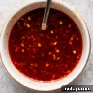 A white bowl filled with a red chili sauce containing visible chili flakes, with a metal spoon resting inside the bowl. The bowl is placed on a textured light gray surface.