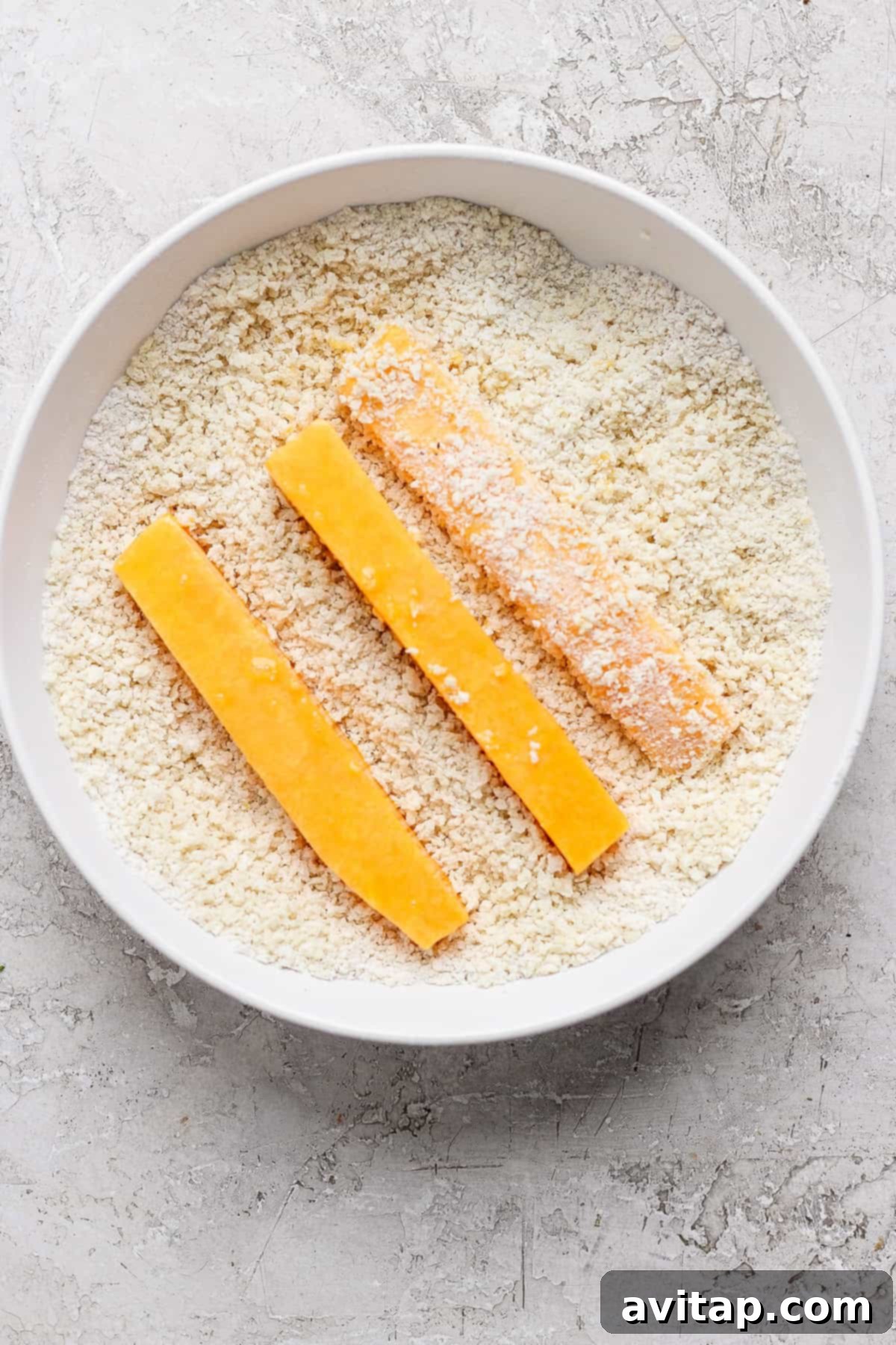 Three breaded cheese sticks are positioned in a bowl filled with breadcrumbs on a textured white surface.