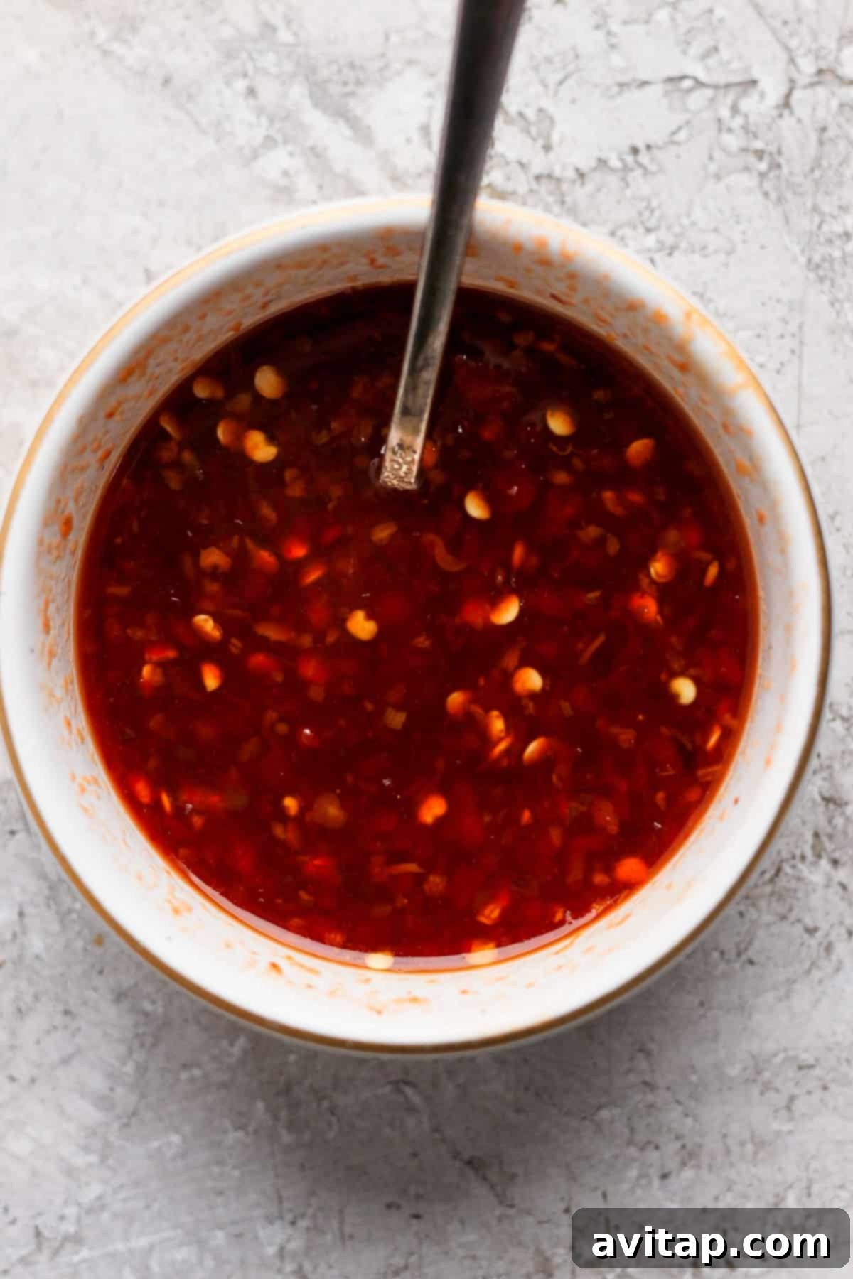 A white bowl filled with a red chili sauce containing visible chili flakes, with a metal spoon resting inside the bowl. The bowl is placed on a textured light gray surface.