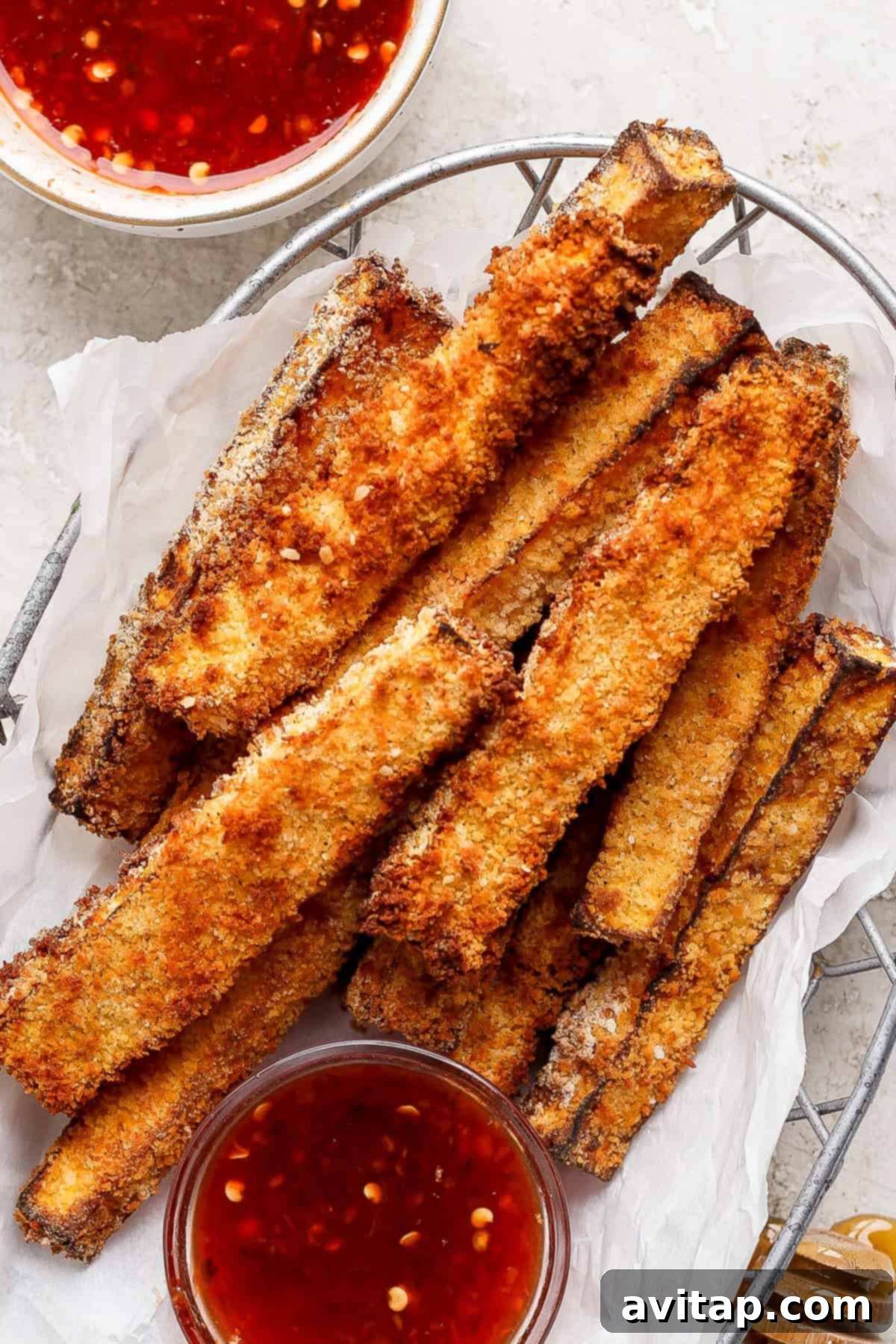 A basket filled with crispy, golden-brown breaded zucchini fries, accompanied by two small bowls of red dipping sauce.