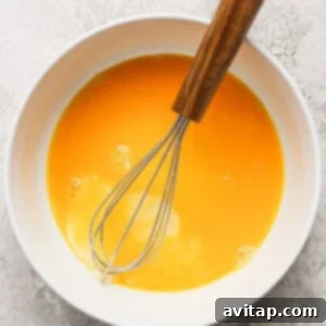 A bowl filled with beaten eggs, with a whisk resting in it on a textured surface.