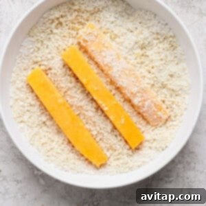 Three breaded cheese sticks are positioned in a bowl filled with breadcrumbs on a textured white surface.