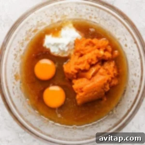 A clear glass bowl containing a mixture of wet ingredients including two raw eggs, pumpkin puree, white yogurt, and a brown liquid, is placed on a textured white surface.