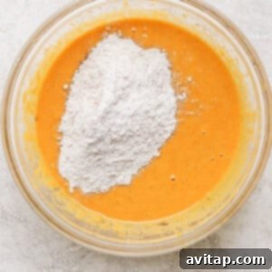 A clear glass bowl containing an orange batter with a heap of white flour added on top, placed on a marble surface, ready for final mixing.
