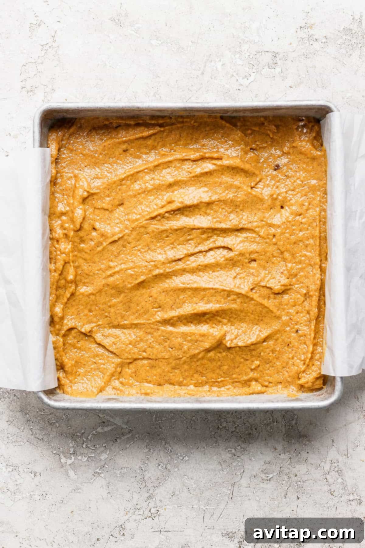 Square baking pan filled with unbaked pumpkin batter on a light textured surface. Edges of the parchment paper extend out from the pan, ready for the streusel.