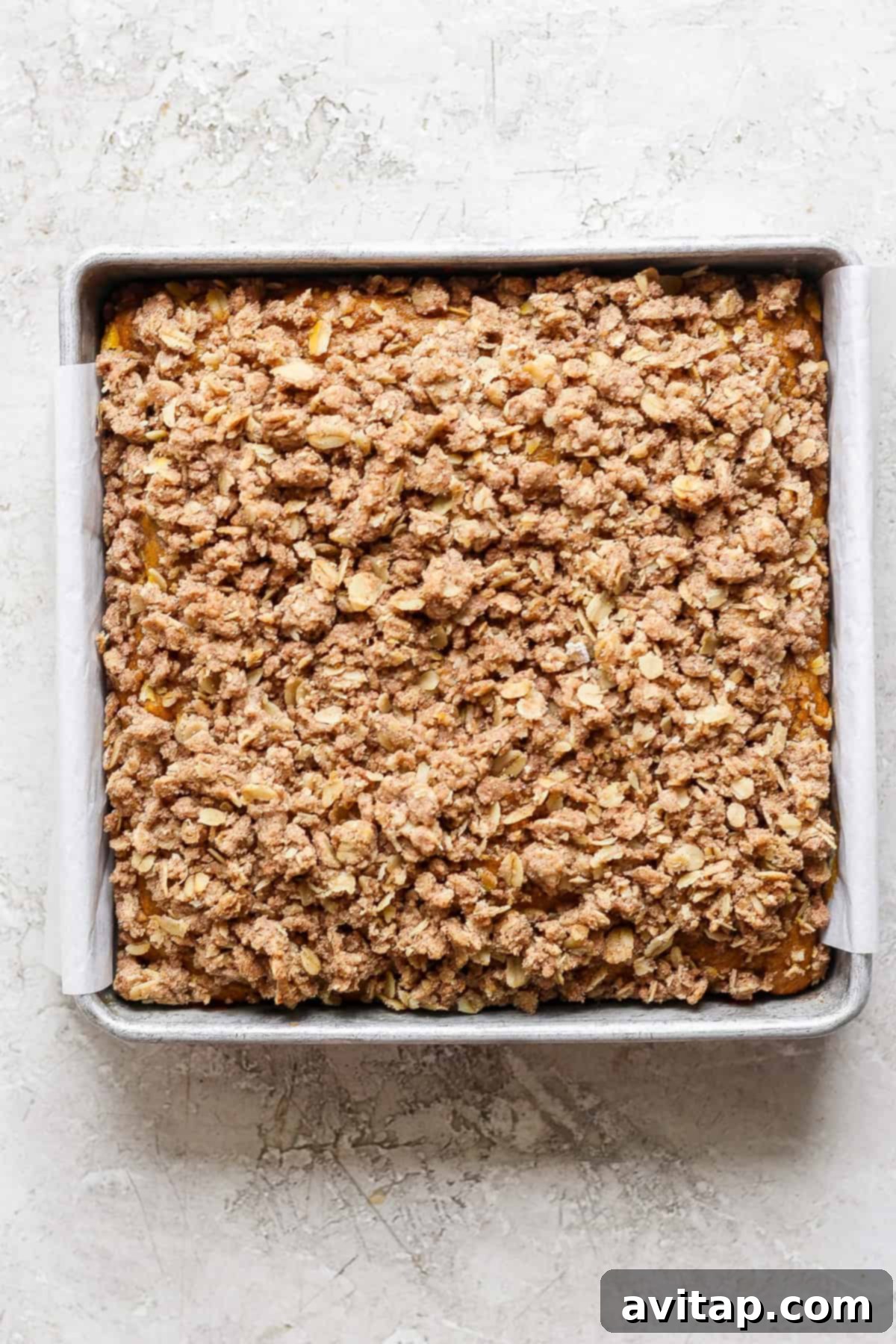 A square metal baking pan filled with a baked oatmeal fruit crumble topped with a crispy, golden-brown oat and cinnamon streusel. The pan is lined with parchment paper, showcasing the perfect crumb.