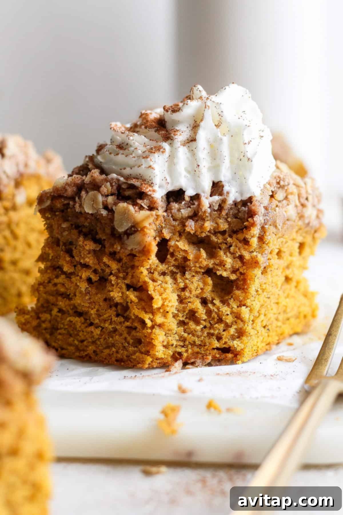 A close-up of a perfectly sliced piece of pumpkin coffee cake, generously topped with streusel and a dollop of whipped cream, presented elegantly on a white surface beside a gold fork.