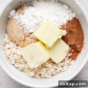 A white bowl containing oats, brown sugar, flour, cinnamon, and two slabs of butter on a light-colored surface, ready for mixing to form streusel.