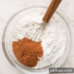 A clear glass bowl containing flour, cinnamon, and a small whisk with a wooden handle on a light-colored surface, ready for dry ingredients.