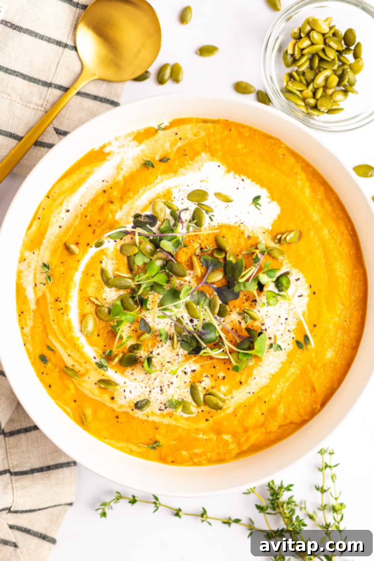 Quick & Creamy Pumpkin Soup 2 A bowl of creamy orange soup garnished with microgreens, pumpkin seeds, and a swirl of cream. A gold spoon, sprigs of fresh herbs, and a small bowl of pumpkin seeds are placed nearby.