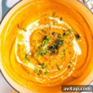 A pot of creamy carrot soup garnished with a swirl of cream, microgreens, pepitas, and black pepper, alongside a piece of bread and herbs.