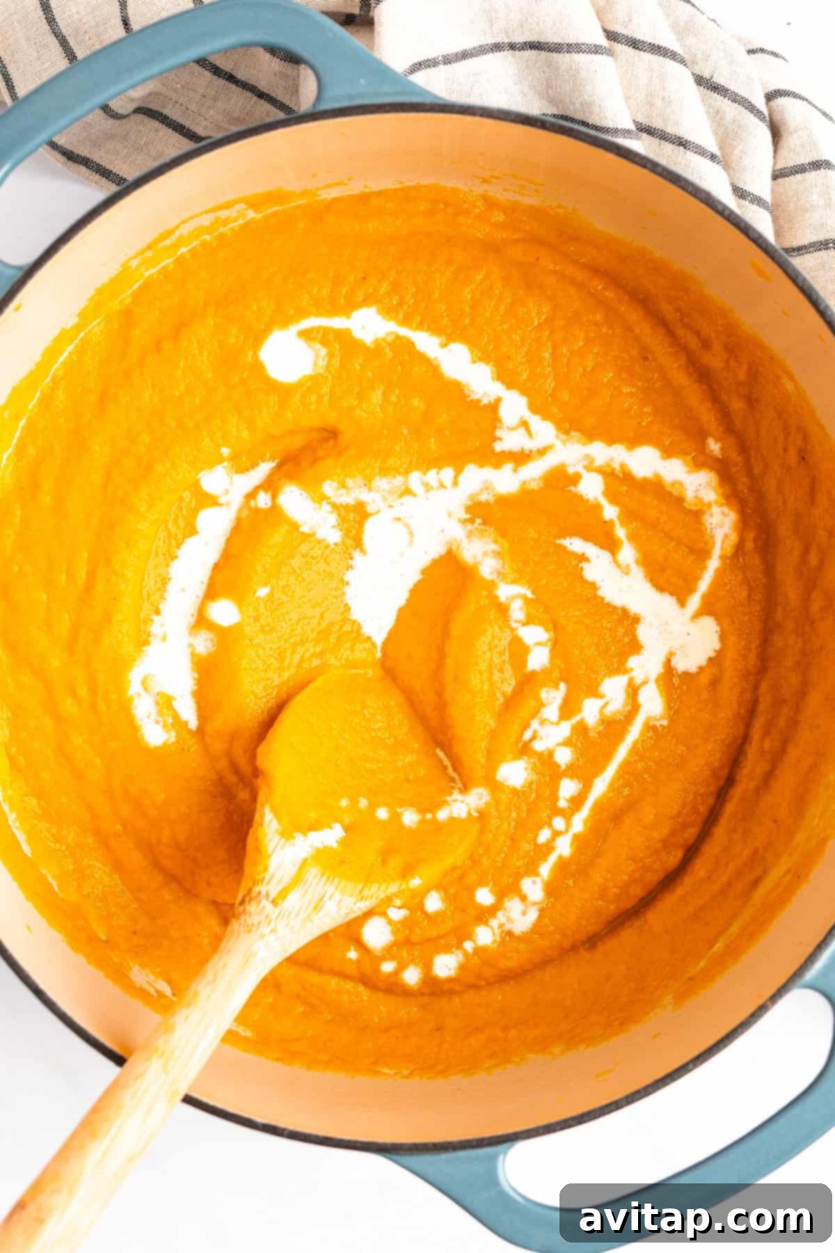 Quick & Creamy Pumpkin Soup 7 A pot of creamy orange soup is being stirred with a wooden spoon. A swirl of cream is visible on top of the soup. A striped cloth is beside the pot.