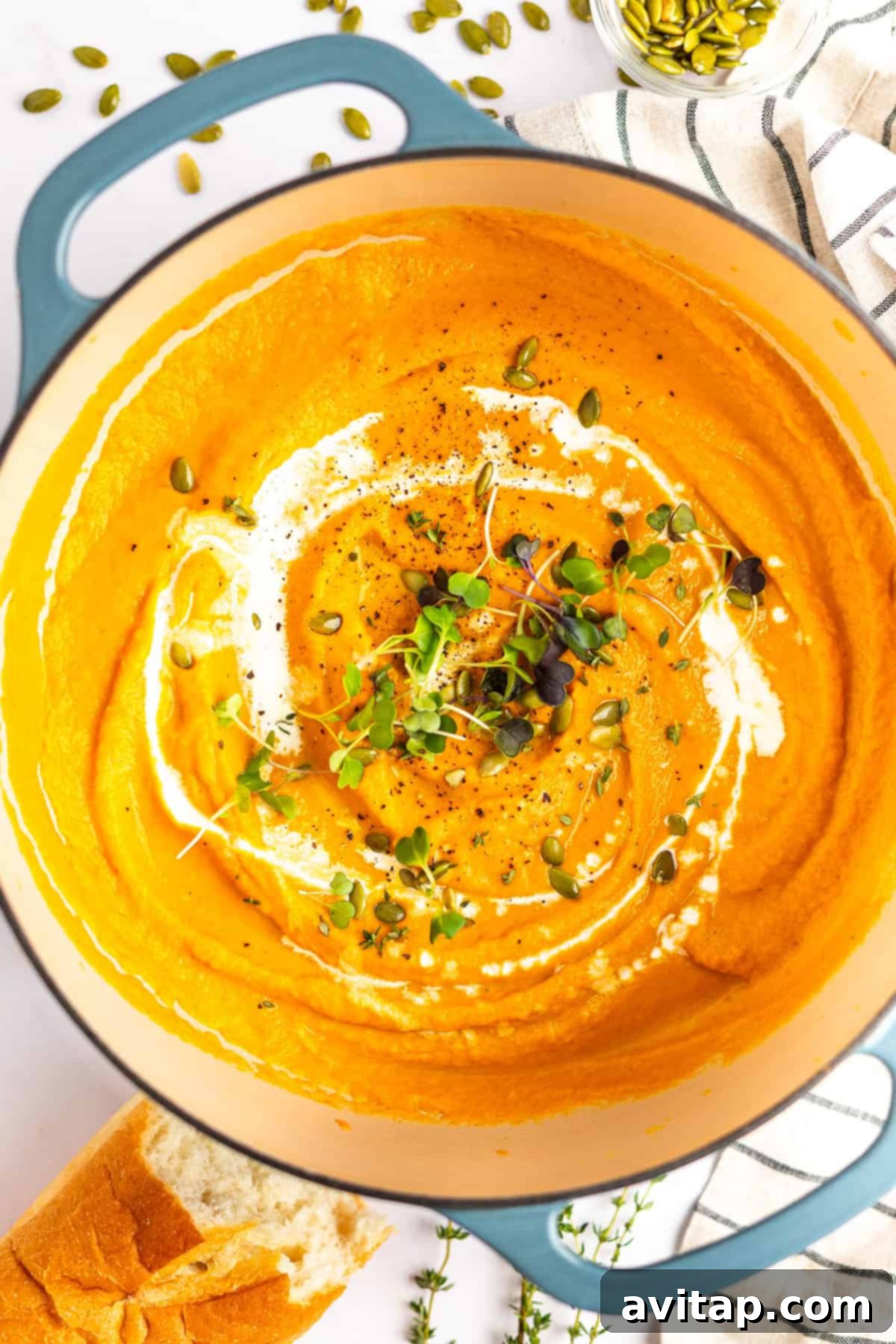 Quick & Creamy Pumpkin Soup 8 A pot of creamy carrot soup garnished with a swirl of cream, microgreens, pepitas, and black pepper, alongside a piece of bread and herbs.