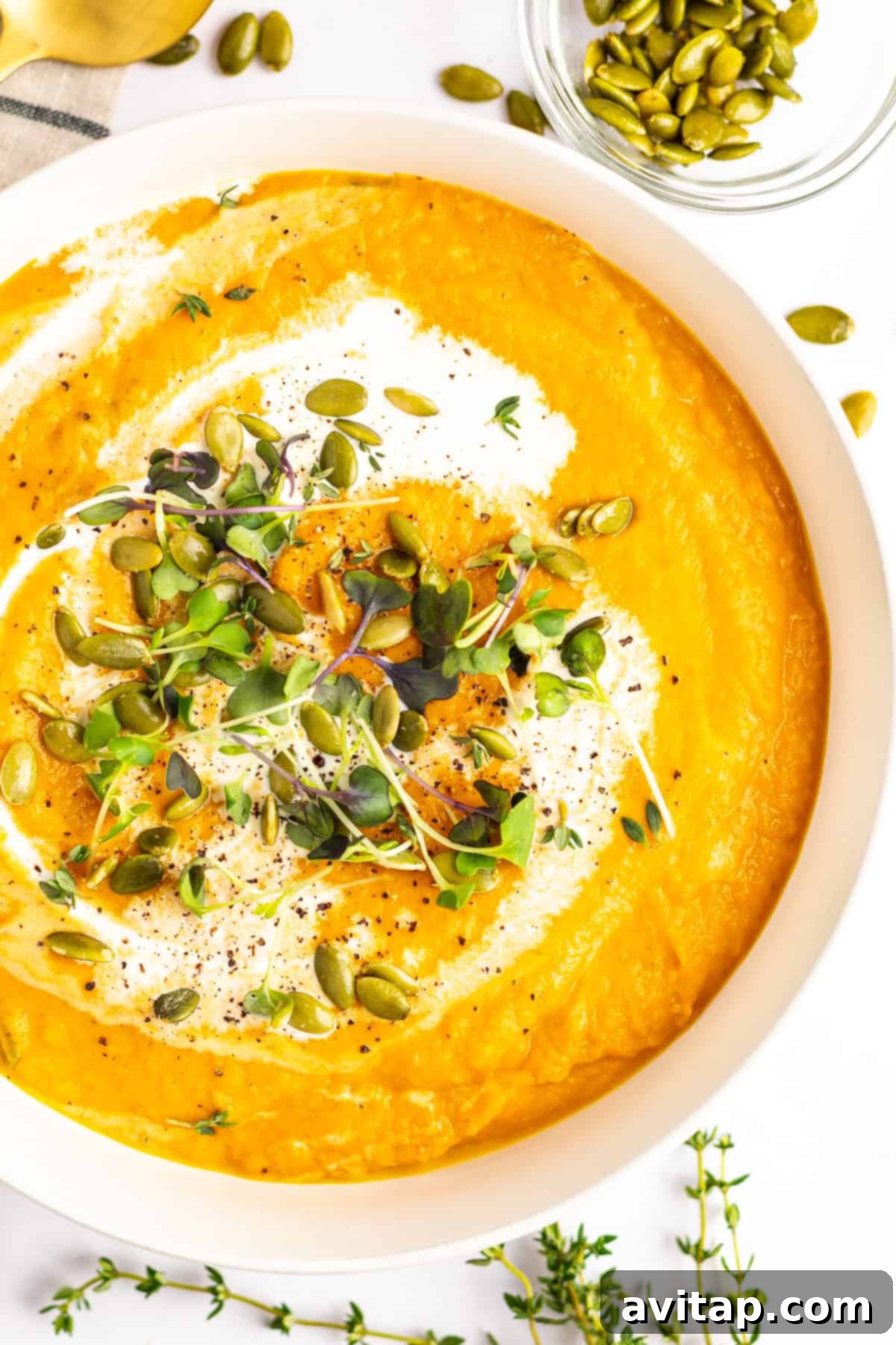 Quick & Creamy Pumpkin Soup 9 A bowl of creamy pumpkin soup garnished with a swirl of cream, microgreens, and pumpkin seeds, with additional pumpkin seeds and thyme sprigs on the side.