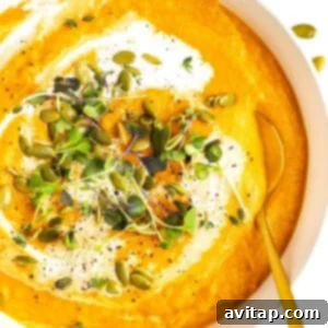 A bowl of creamy orange soup garnished with fresh microgreens, pumpkin seeds, black pepper, and a swirl of cream. A golden spoon rests inside the bowl.