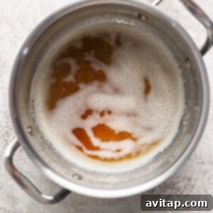 A metal pot with handles containing a frothy liquid that appears to be browning. The pot is placed on a light, textured surface.