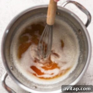A hand holding a whisk stirs frothy liquid with brown patches in a stainless steel pot.
