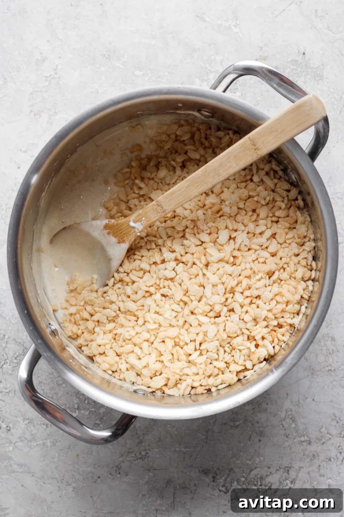 A pot containing a mixture of melted marshmallows and brown butter, with rice cereal being stirred in with a wooden spoon on a gray surface.