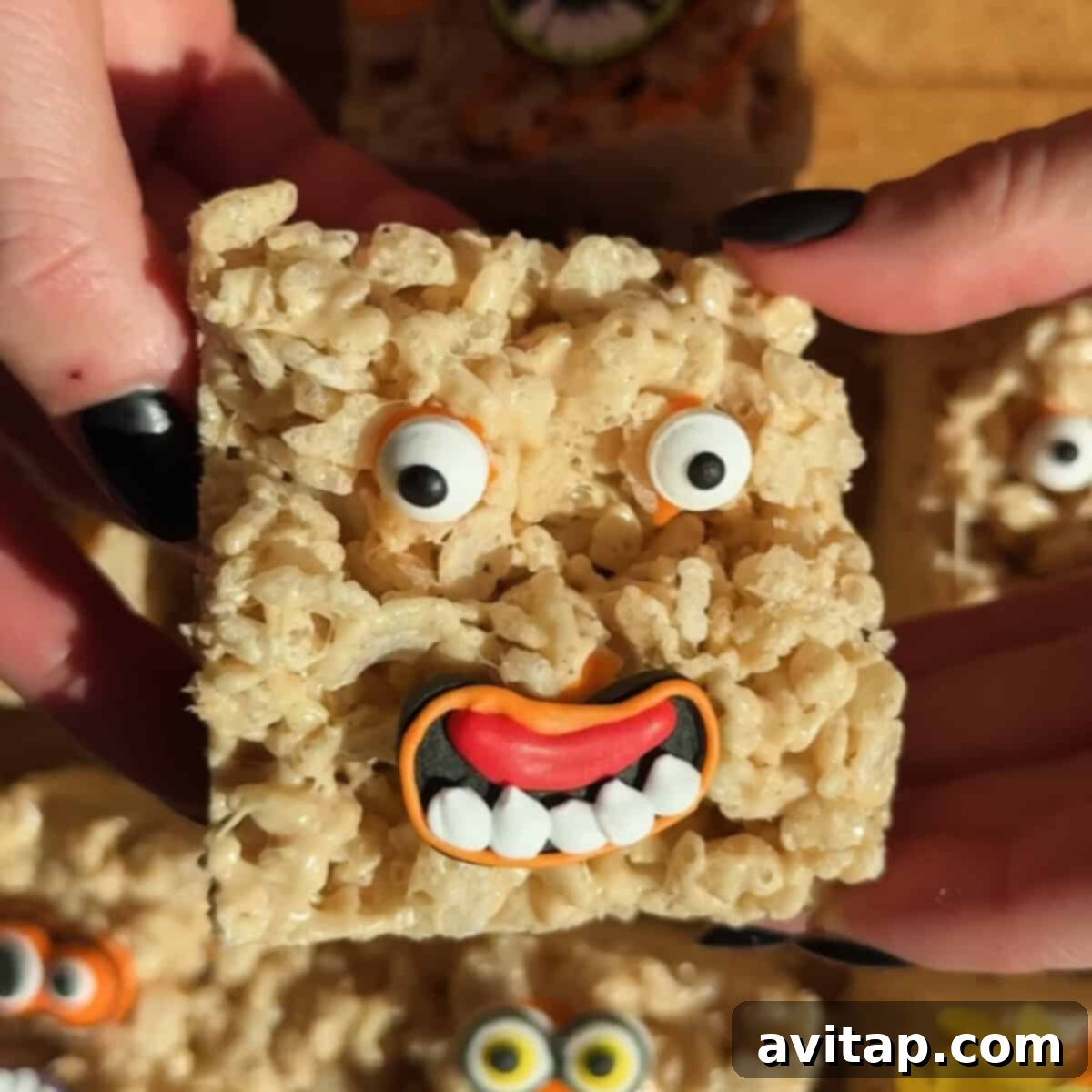 A Rice Krispies treat decorated with candy eyes and a friendly mouth, resembling a goofy face, is held between two hands with black-painted nails.
