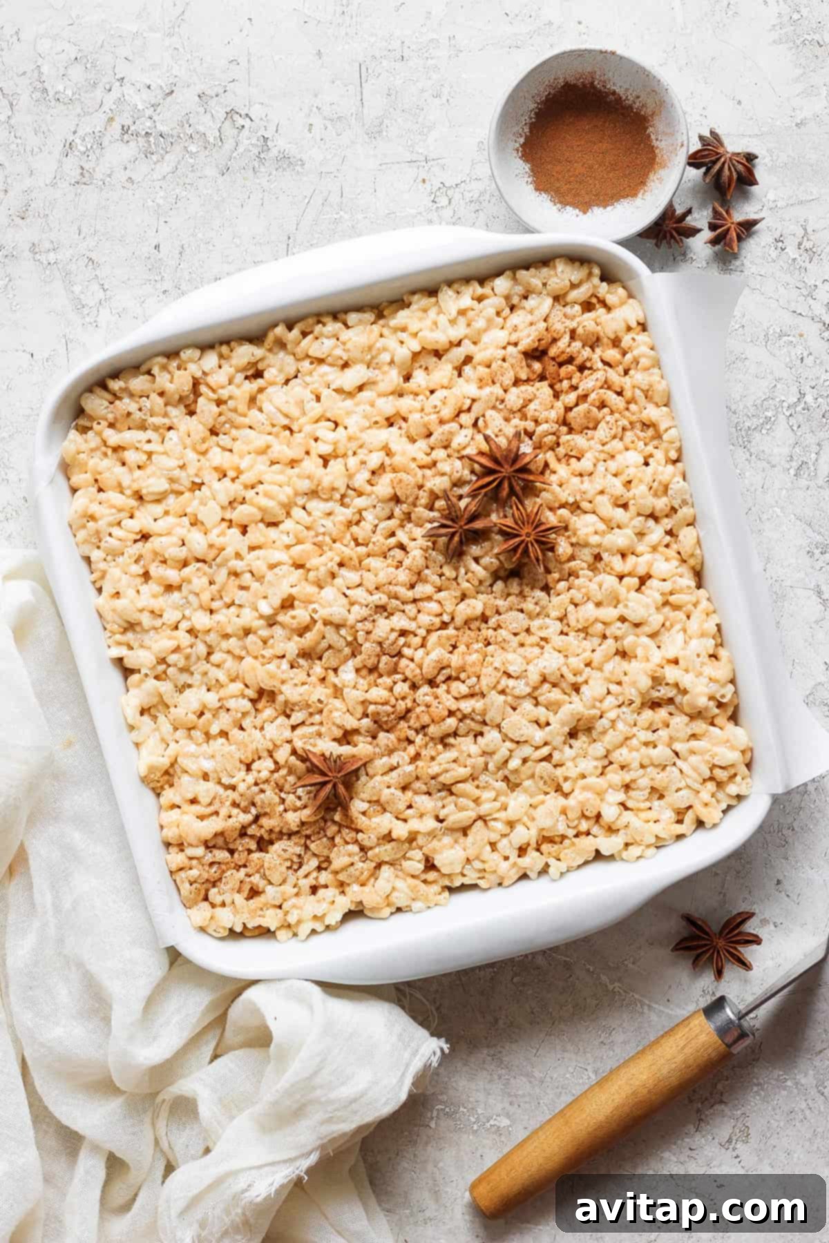 A square white baking dish filled with rice crispy treats, garnished with star anise and cinnamon powder in the top right corner. A small bowl with cinnamon powder is beside the dish on the counter, ready for dusting.