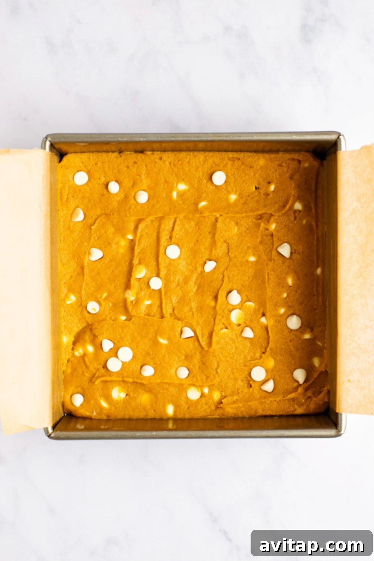 Pumpkin Spice Blondies 3 A square baking pan filled with unbaked batter topped with scattered white chocolate chips, lined with parchment paper on a marble countertop.