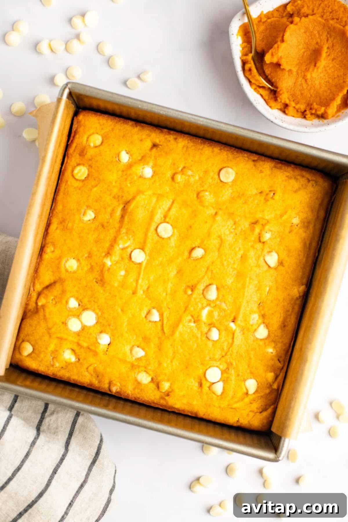 Pumpkin Spice Blondies 4 A square pan of orange-colored baked goods with white chocolate chips. A small bowl of pumpkin puree and a spoon are placed beside the pan. A striped cloth and scattered chocolate chips are also visible.