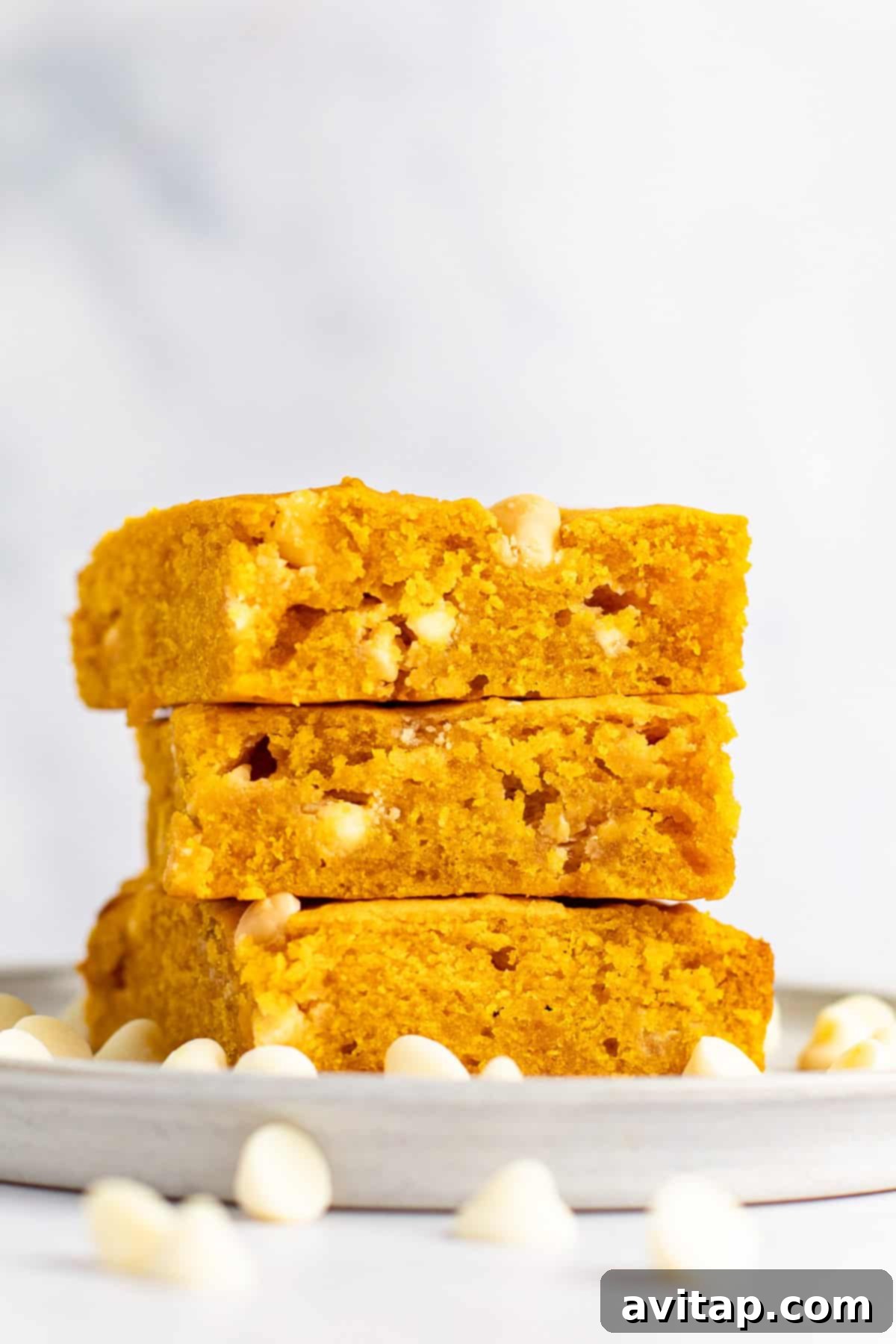 Pumpkin Spice Blondies 5 A stack of three pumpkin-flavored blondies with white chocolate chips sits on a grey plate, surrounded by scattered white chocolate chips, against a white background.