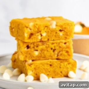 Pumpkin Spice Blondies 6 A stack of three pumpkin blondies with white chocolate chips on a white plate. Scattered white chocolate chips surround the stack.