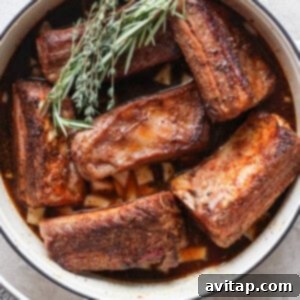 A white pot contains several pieces of cooked beef short ribs garnished with fresh rosemary and thyme.
