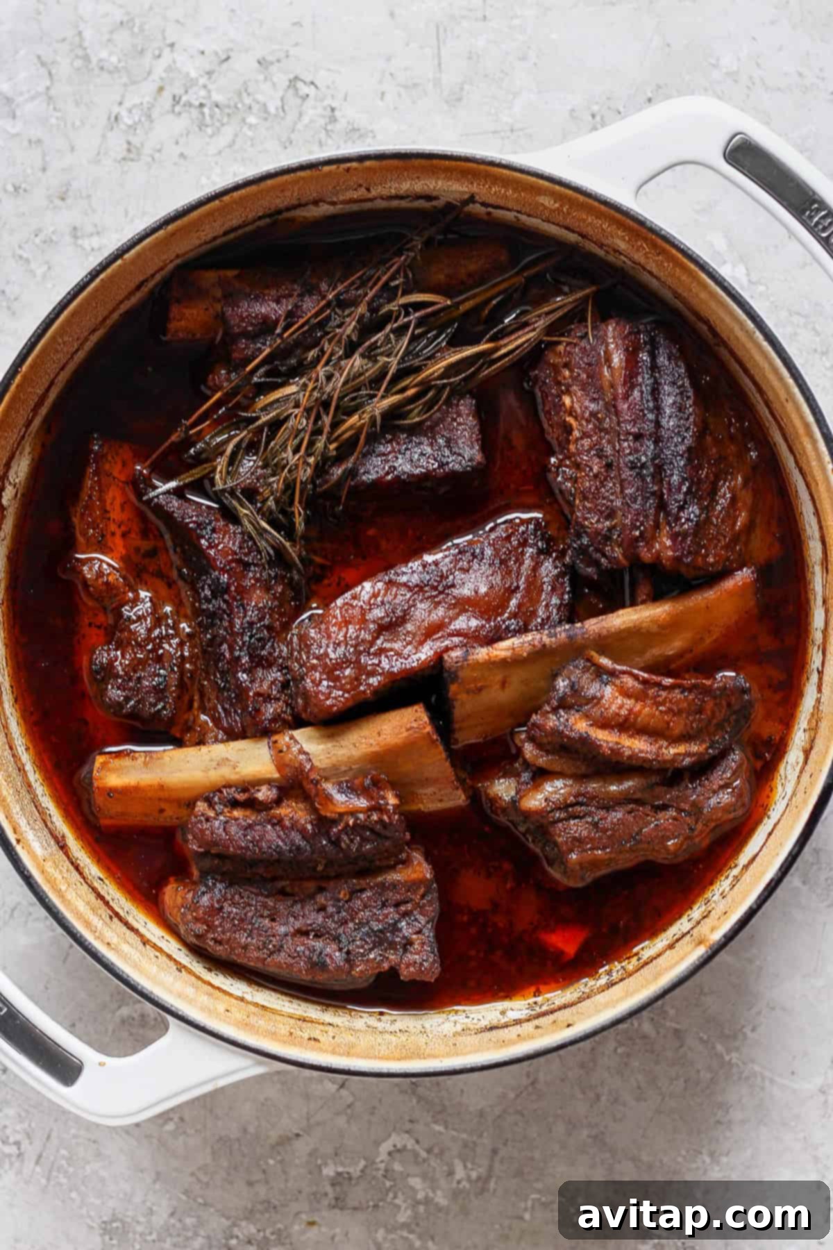 A large pot filled with braised short ribs in a rich, dark sauce, garnished with herbs on a light gray surface.