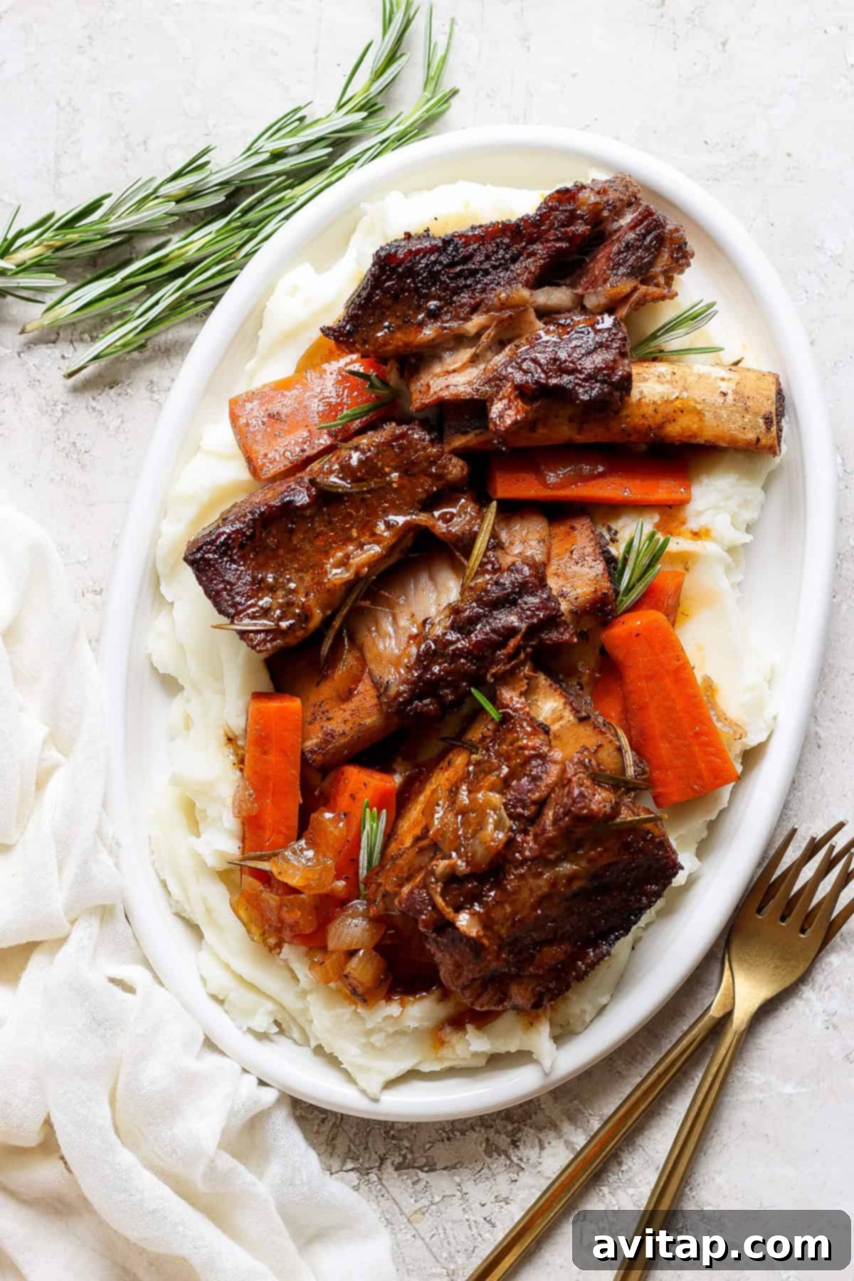A white oval plate with mashed potatoes, roasted carrot slices, and beef short ribs garnished with fresh rosemary sprigs. A fork and knife are placed beside the plate on a white surface.