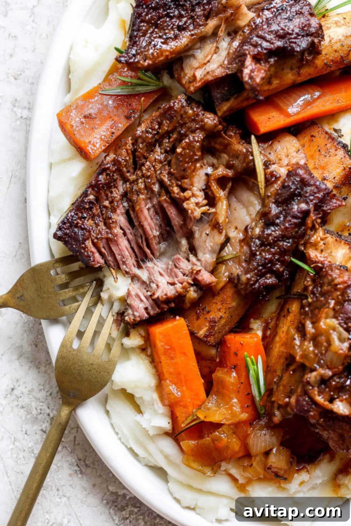 A plate of tender braised beef short ribs with carrots, garnished with rosemary, served on a bed of creamy mashed potatoes. Two forks are placed beside the dish.