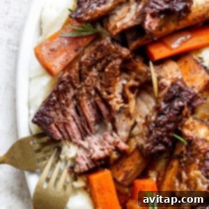 A plate of tender roasted meat with a crispy exterior, served on a bed of mashed potatoes and garnished with cooked carrots and rosemary, with two forks visible on the side.