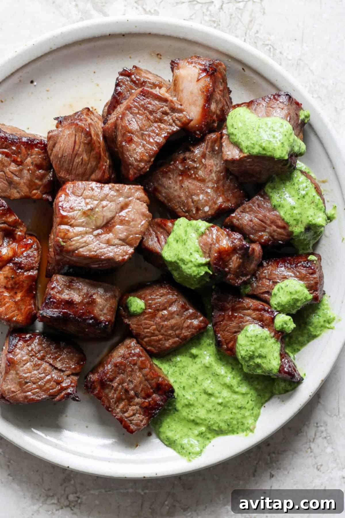A close-up shot of a white plate featuring expertly grilled steak bites, lavishly topped with a vibrant green herb sauce.